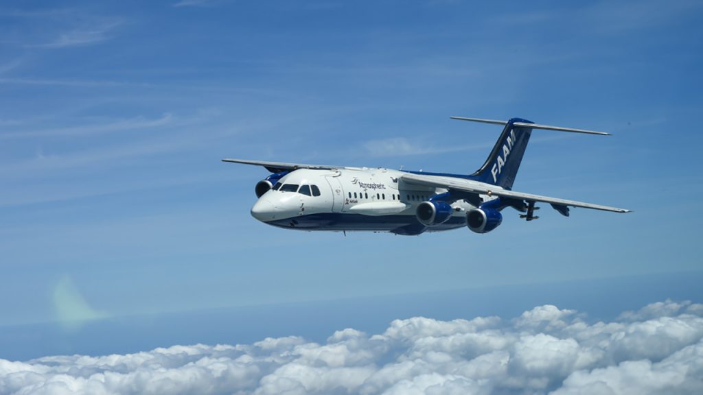 A white and blue aircraft flies above clouds in a blue sky