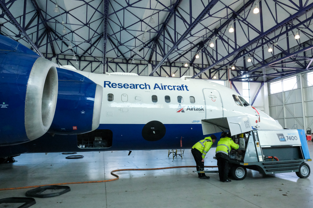 Two people wearing hi vis jacket bend over looking inside a metal unit on wheels, beside a blue and white aircraft