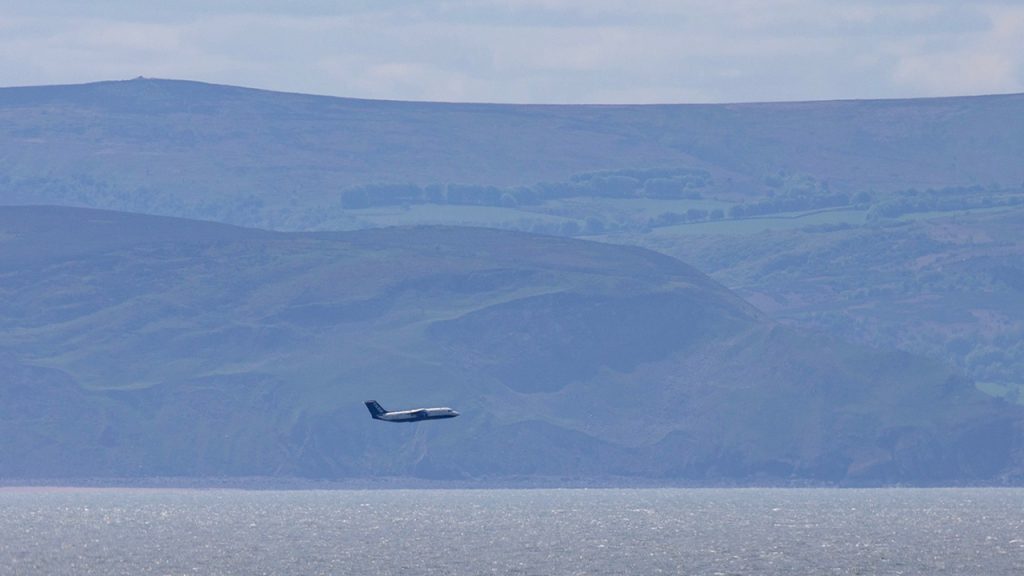 An atmospheric research aircraft flying at a low level over the sea, with hills in the background.