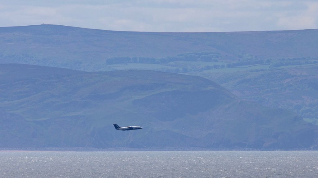 An atmospheric research aircraft flying at a low level over the sea, with hills in the background.