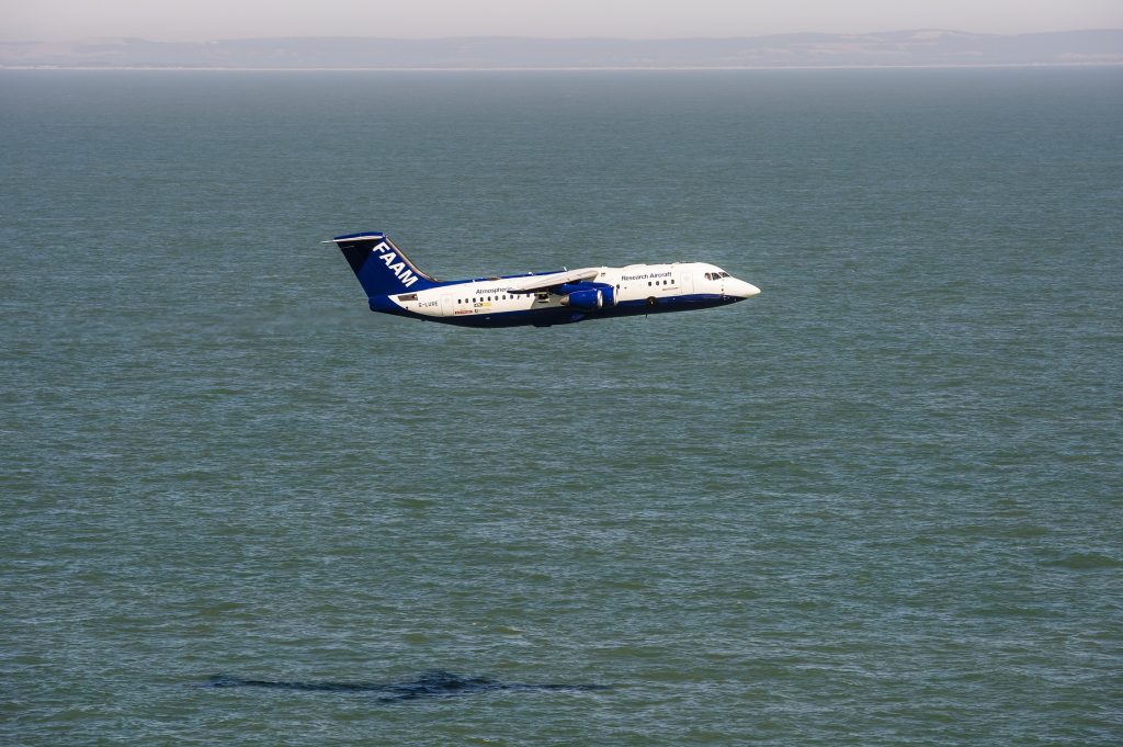 A blue and white atmospheric research aircraft flying at low level over a calm sea