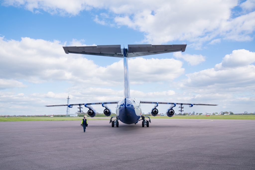 Blue and white research aircraft parked on apron. Person wearing high visibility jacket walks away from aircraft. Green grass and blue sky with clouds in the background.