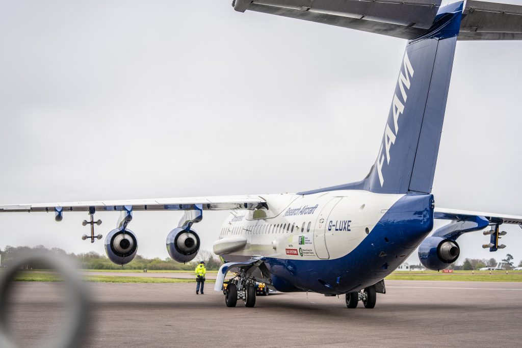 Blue and white research aircraft being towed across apron with person wearing high visibility jacket walking next to it.