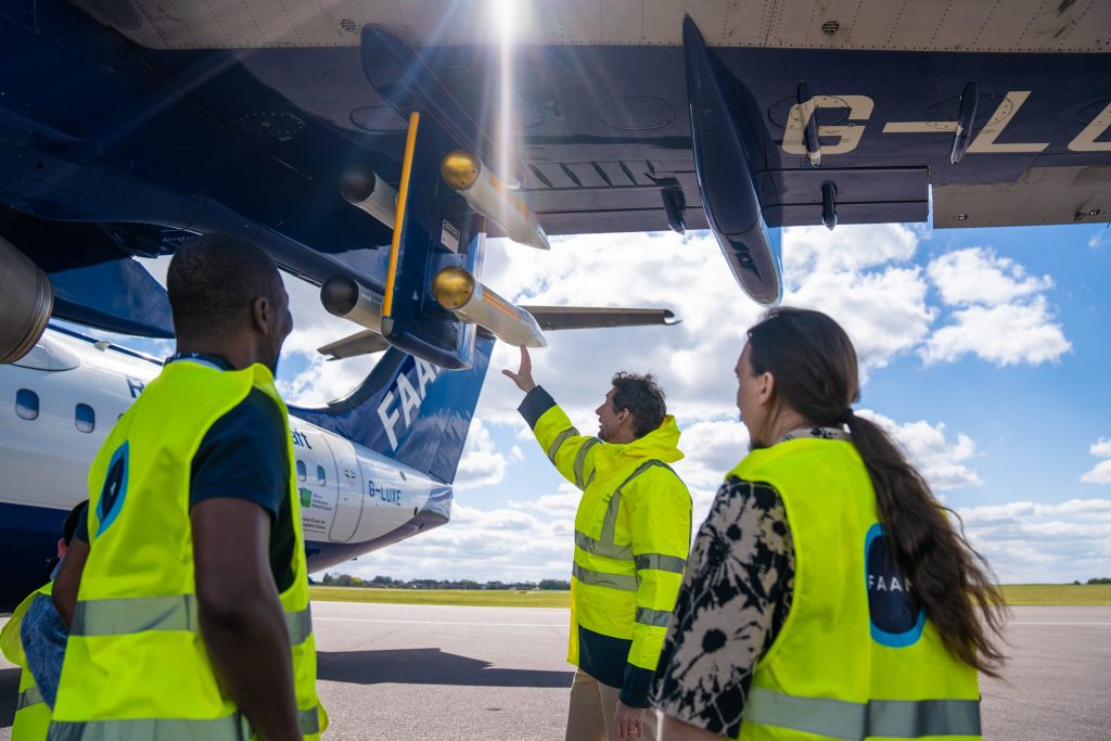 Four people wearing high visibility jackets look at the instrument pylon on the wing of a blue and white research aircraft. One person is pointing towards the aircraft.