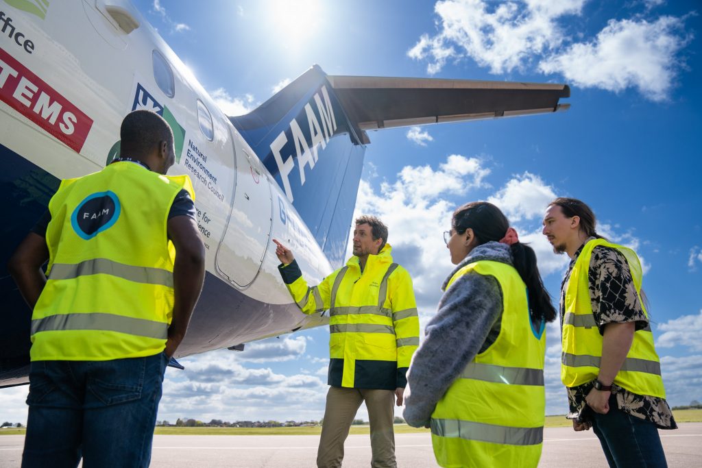 Four people wearing high visibility jackets look at blue and white research aircraft. One person is pointing towards the aircraft.