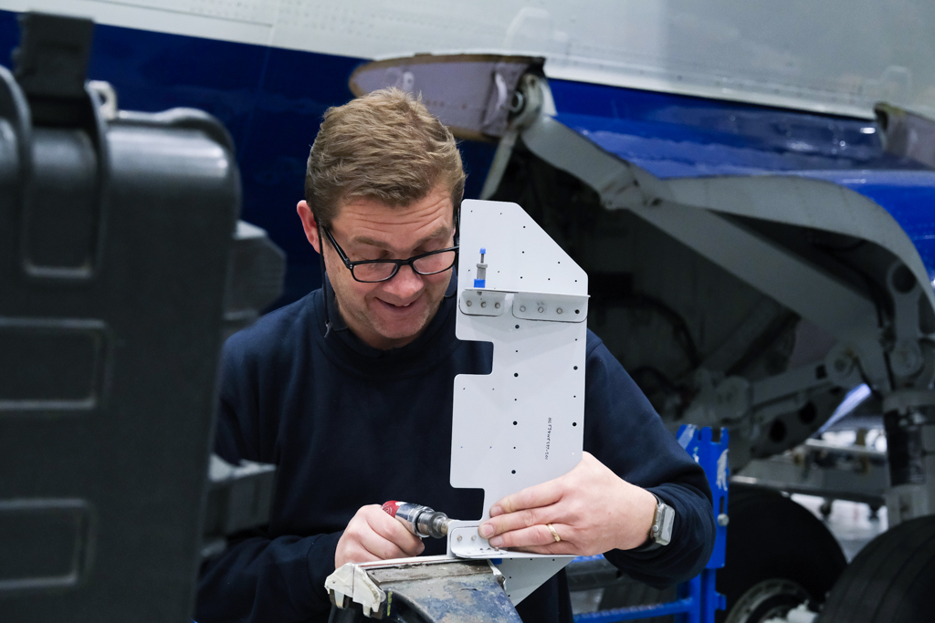 A person uses a tool to modify a metal bracket. They are standing in front of a large research aircraft.