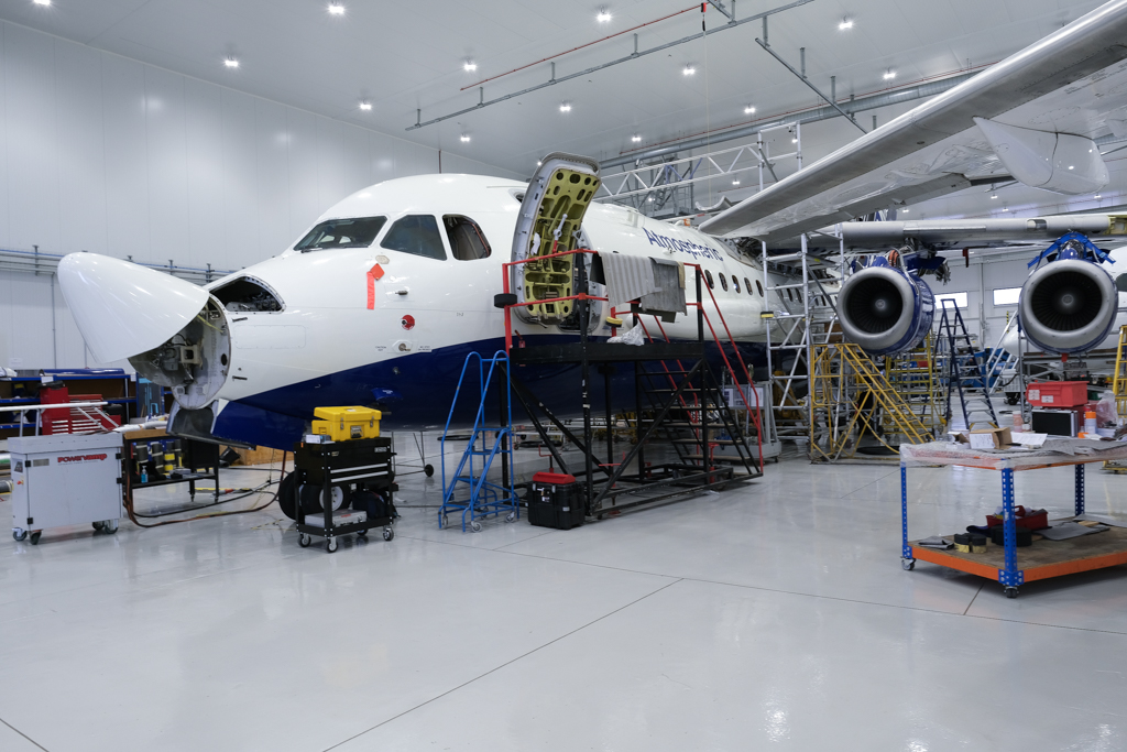 A large blue and white research aircraft in a hangar. It has scaffolding around is and is undergoing maintenance, with the nose cone flipped up and several exterior panels removed.