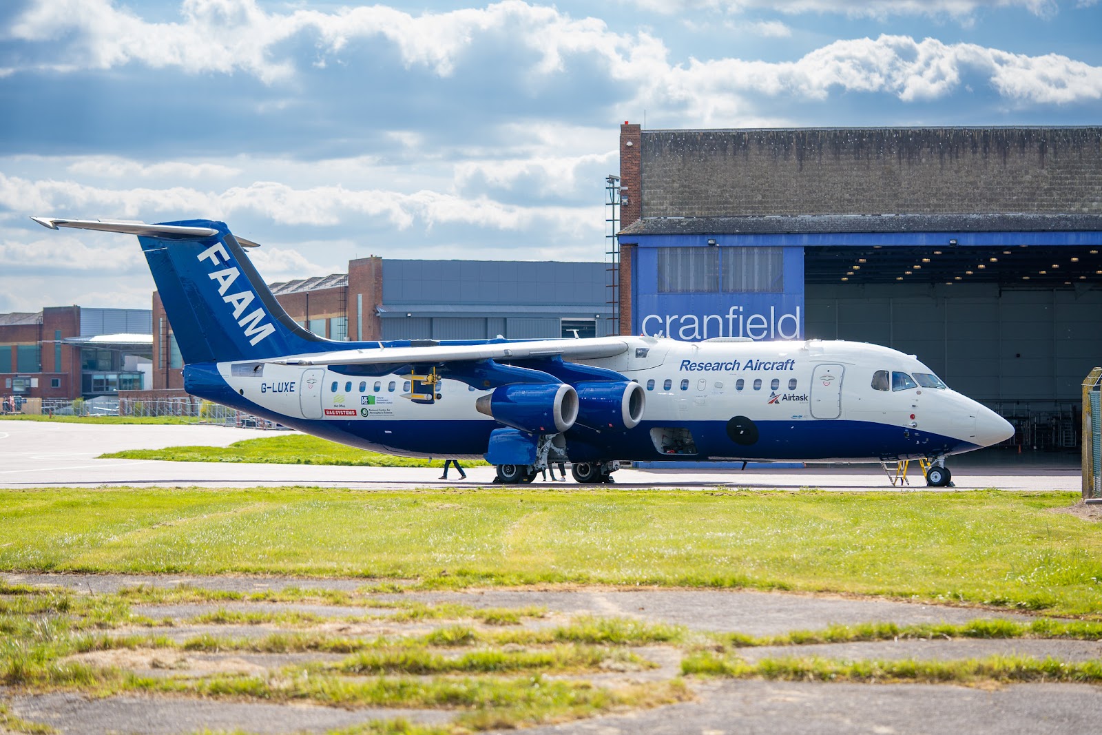 A large blue and white atmospheric research aircraft parked outside a hangar in sunshine.