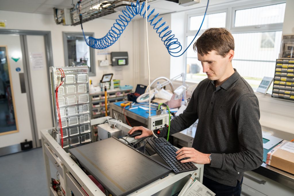 Person inside laboratory stood at an instrument rack, typing on a keyboard and looking at a computer screen.