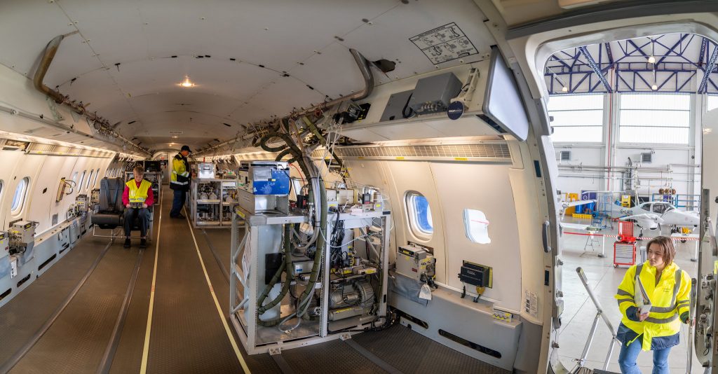 Three people inside research aircraft. One person sat in a seat looking at a screen. Another person stood looking at equipment. Third person is climbing stairs into aircraft.