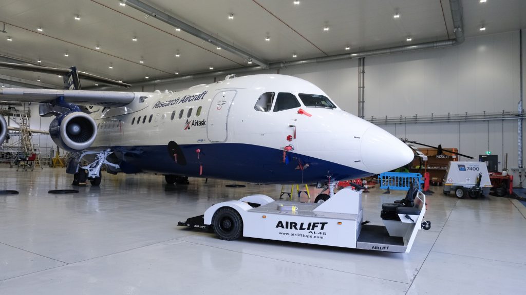 A white electric towing vehicle parked in front of a large blue and white research aircraft inside a hangar.