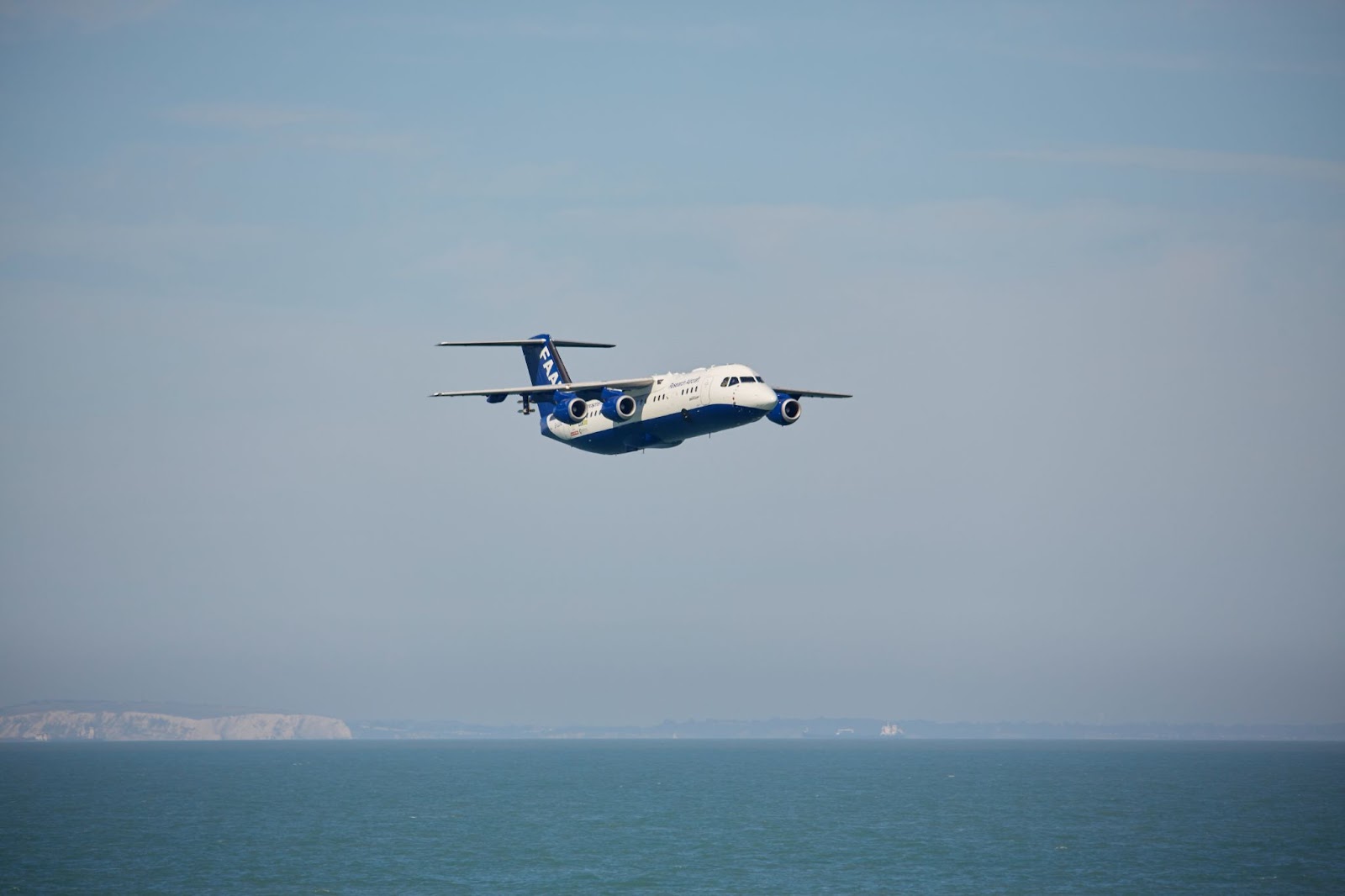 A large blue and white research aircraft flying low over the sea. White cliffs are visible on the horizon.