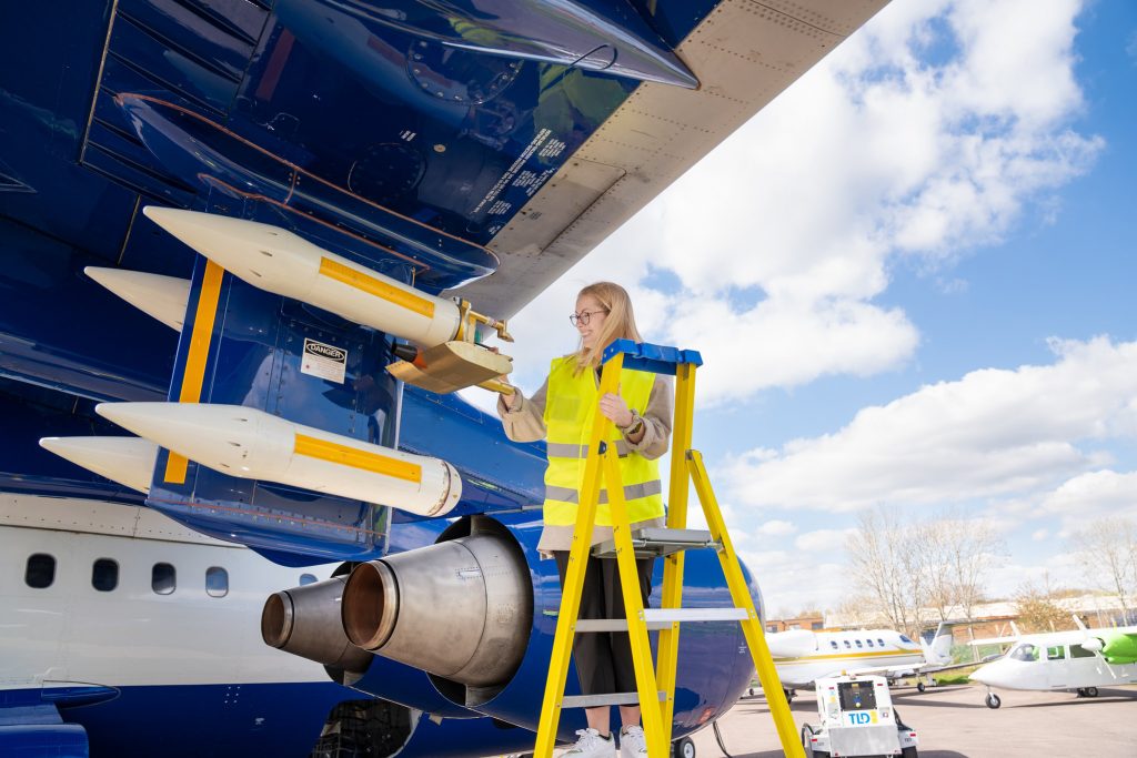 Person wearing a high visibility jacket stood on step ladder. Person is inspecting instruments on wing of blue and white research aircraft.