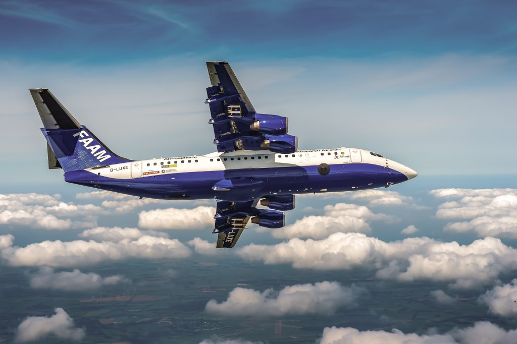A large blue and white research aircraft banking away from the camera as it flies above puffy white clouds.