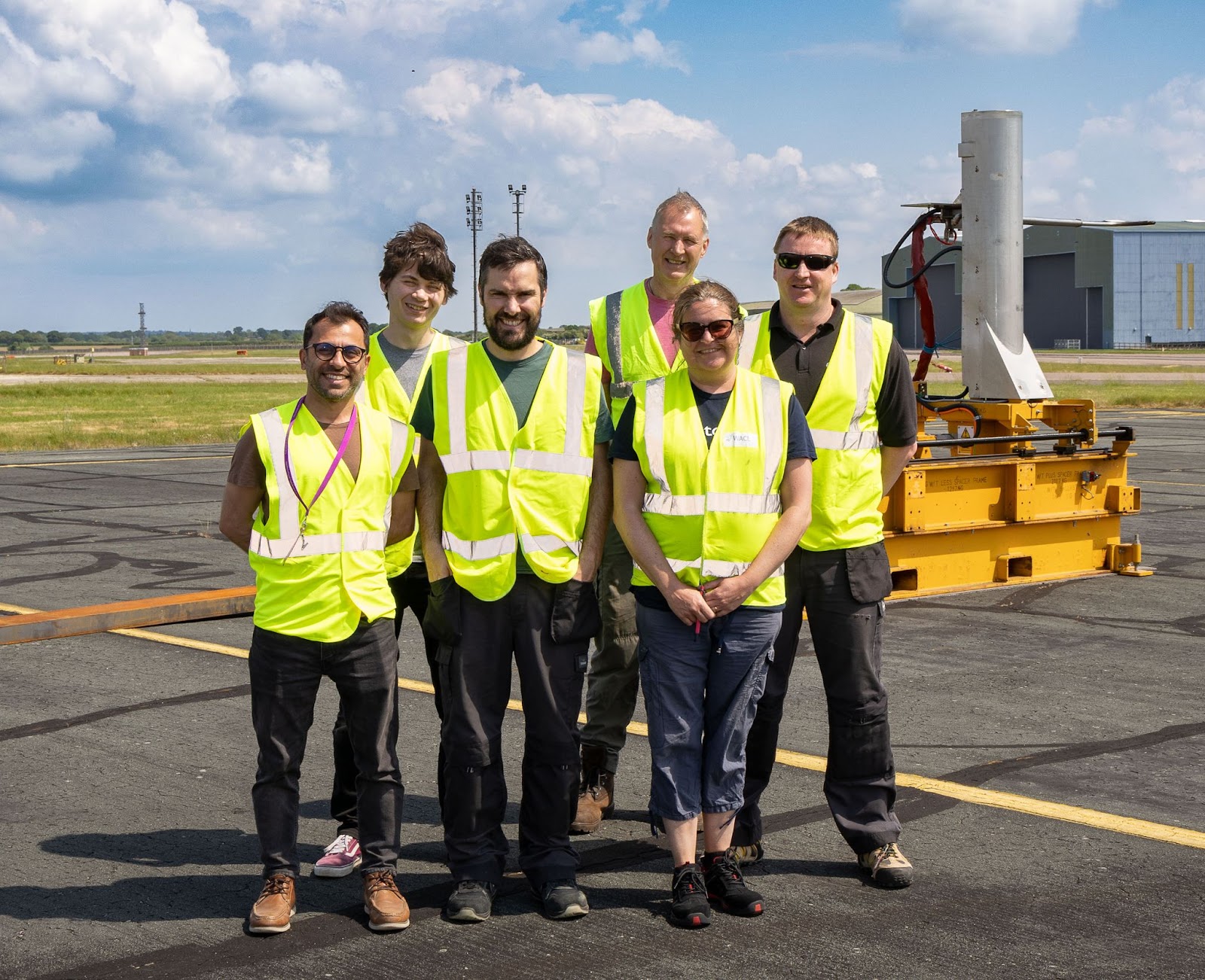 Six people in hi-vis jackets in front of a large piece of emissions measuring equipment at an airport. They are smiling at the camera.